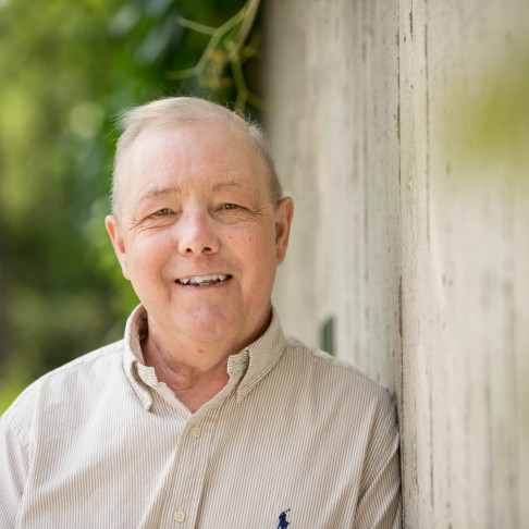 David, a middle-aged man, looking at camera, smiling, while leaning against a concrete wall with lush greenery filling the background.