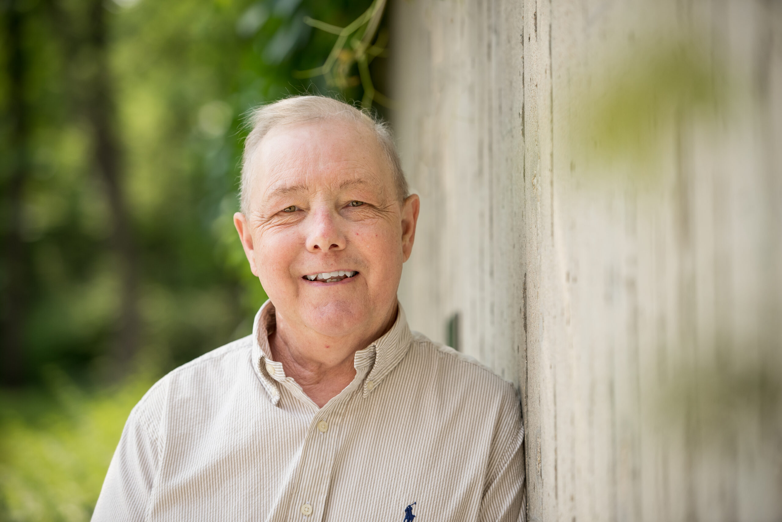 David, a middle-aged man, looking at camera, smiling, while leaning against a concrete wall with lush greenery filling the background. 