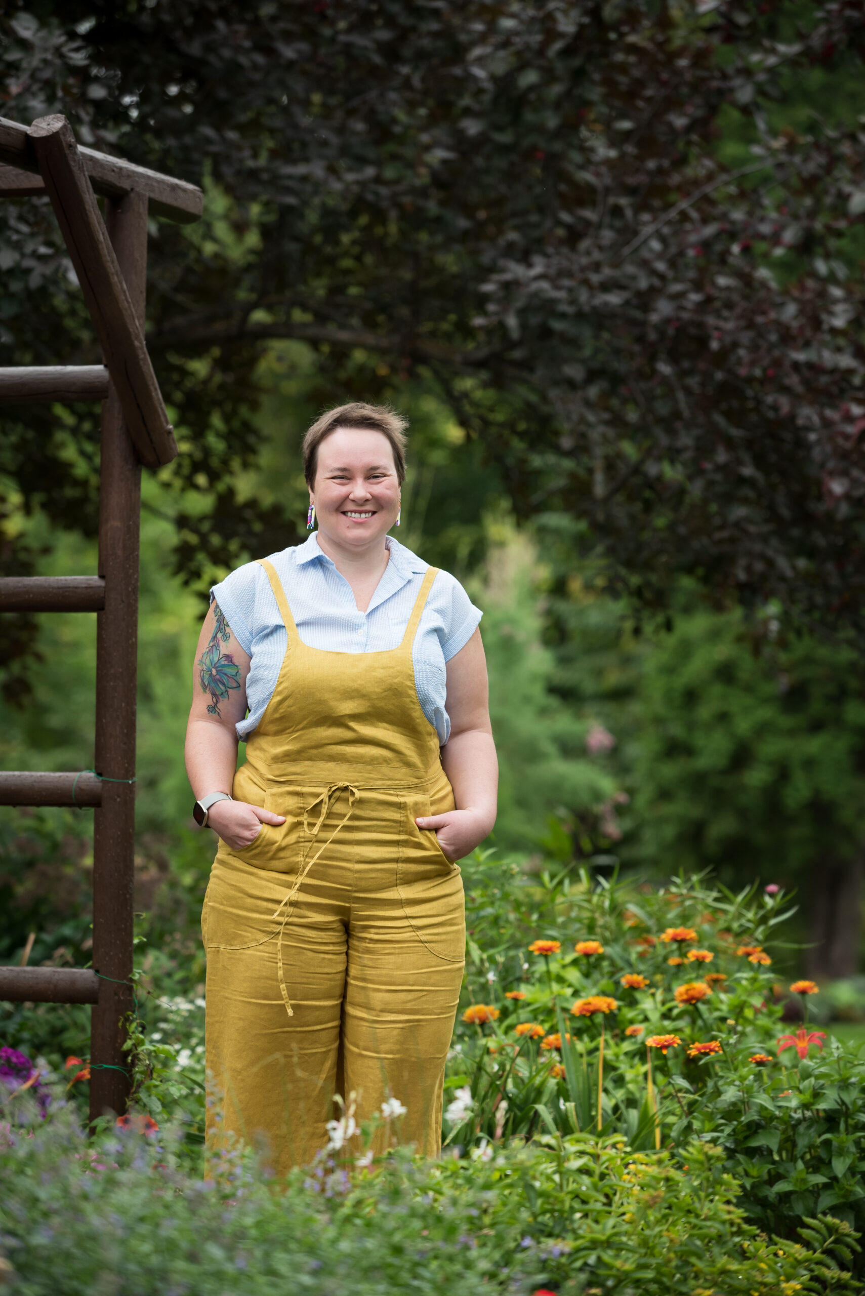 Young woman in her thirties wearing a yellow jumper with a light blue collared shirt posing for a photo in a park setting by orange flowers, brown stained wooden arbour.