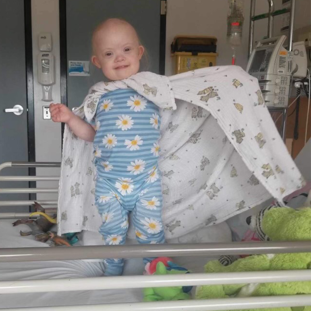 Ruby in pajamas in hospital room with blanket wrapped around her like a cape.